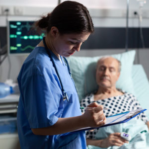 Medical assistant checking treatment of senior man reading notes on clipboard. Sick elderly laying in bed discussing with nurse about diagnosis expertise with iv bag attached.